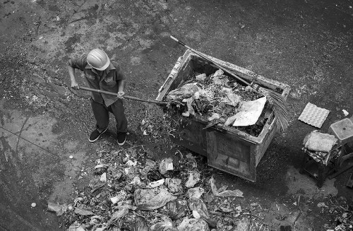 A person wearing a safety helmet and casual clothing is using a long-handled tool to clear loose rubbish from the ground near a large industrial skip, which is filled with mixed waste materials, including paper, plastic, and other debris. The scene takes place outdoors on a paved or concrete surface, with scattered waste around the base of the skip. The skip itself is positioned along a roadside or driveway, with visible urban surroundings. The individual is standing on the left side of the image, leaning slightly forward as they work, with the waste spilling onto the ground beneath. The area appears to be part of a waste collection or rubbish disposal site, with an emphasis on rubbish removal services that might be offered by companies like Office Clearance East Ham, operating within East Ham or nearby postcode areas. The image's black-and-white tone highlights the textures of the waste, the rough surface of the ground, and the utilitarian environment involved in waste collection and disposal processes.