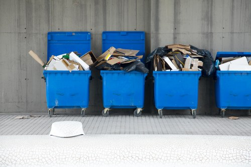 A row of five bright blue wheeled rubbish bins aligned along a grey concrete wall on a paved surface, each filled with various types of discarded waste materials. The leftmost bin contains a mix of white and green packaging, with some cardboard and plastic waste visible. The second bin holds cardboard boxes, some flattened and others partially collapsed, with brown and beige tones. The middle bin appears to contain a large black plastic bag filled with rubbish, and some loose cardboard pieces spilling out. The fourth bin is filled with more cardboard and paper waste, with some protruding over the top edge. The rightmost bin also contains assorted waste, including packaging and paper. A single white waste item, possibly a paper plate, rests on the ground in front of the bins, and the setting suggests an area designated for rubbish disposal outside a commercial or residential building in East Ham, London, capturing a typical scene of waste awaiting removal as part of a rubbish clearance service by Office Clearance East Ham.