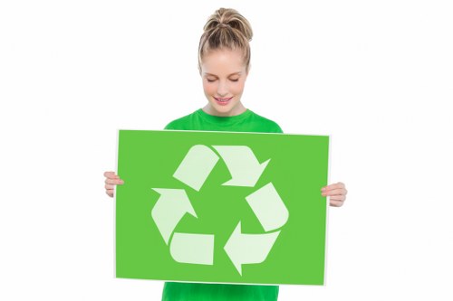 A young woman with light brown hair tied in a ponytail, wearing a bright green long-sleeved shirt, is smiling gently while holding a large rectangular green sign featuring a white recycling symbol composed of three arrows forming a triangle. The background is plain white, emphasizing the woman's cheerful expression and the prominent recycling graphic. This image reflects environmentally conscious messaging and eco-friendly practices, relevant to waste management and rubbish removal services often associated with companies like Office Clearance East Ham. The woman’s casual attire and the vivid green color of the sign symbolize sustainability and responsible disposal, aligning with the themes of rubbish collection and waste recycling typically provided in East Ham and surrounding areas.