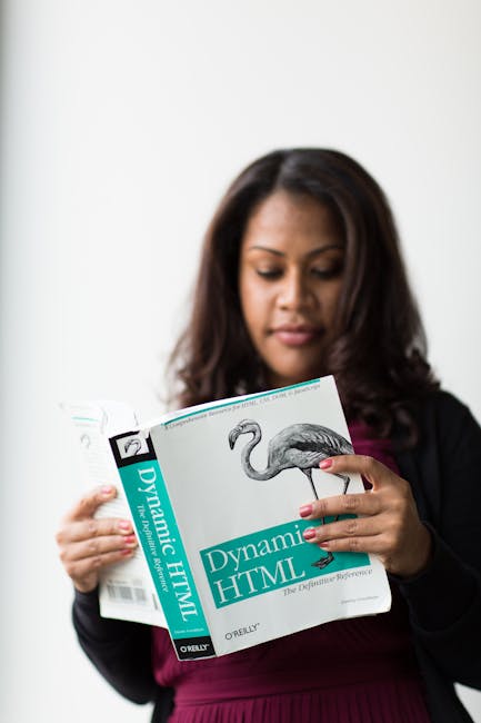 A woman with dark, shoulder-length hair and medium skin tone is standing indoors against a plain white wall. She is wearing a black blazer over a burgundy top and is focused on reading a magazine titl