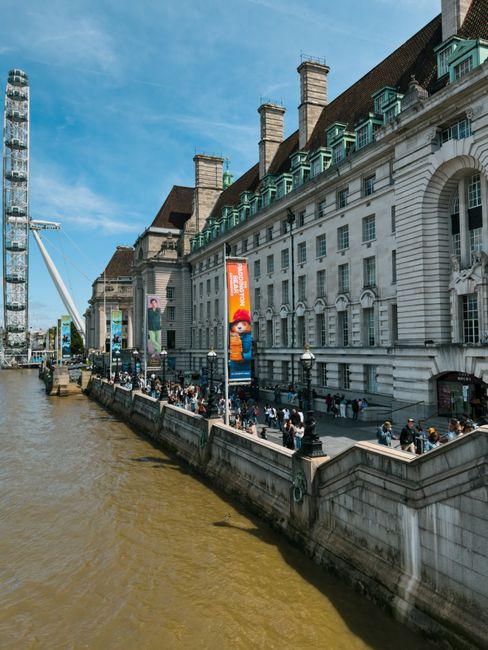 A historic white stone building with multiple chimneys and a dark tiled roof is situated along the riverbank, with several tall, colorful banners hanging from its facade. The building features arched 