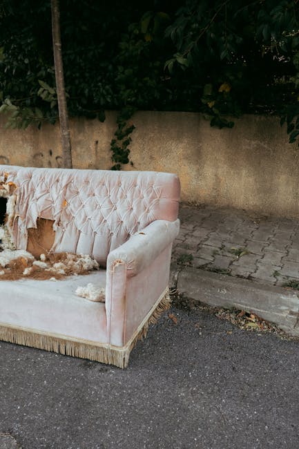 A large, three-seater upholstered sofa is positioned outdoors against a brick wall backdrop. The sofa has a fabric covering in a textured, medium-grey tone with visible button tufting on the backrests